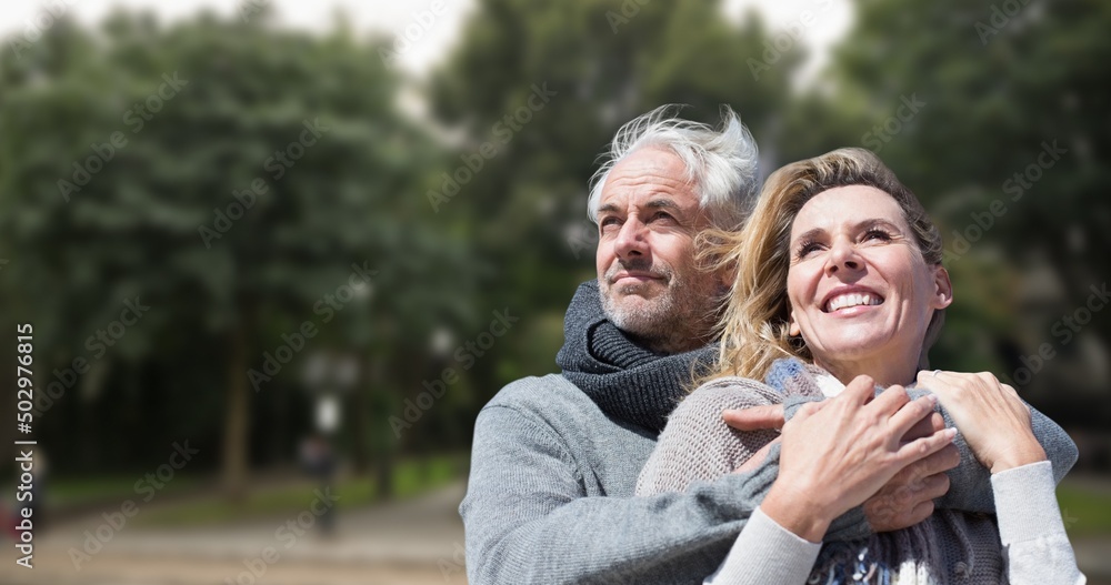 Smiling caucasian senior man embracing senior woman against trees in park during sunny day