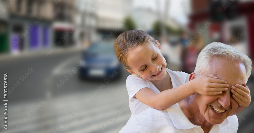 Smiling playful caucasian granddaughter covering grandfathers eyes against street in city