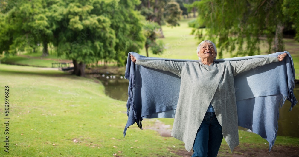 Cheerful caucasian senior woman holding scarf with arms outstretched against trees in park