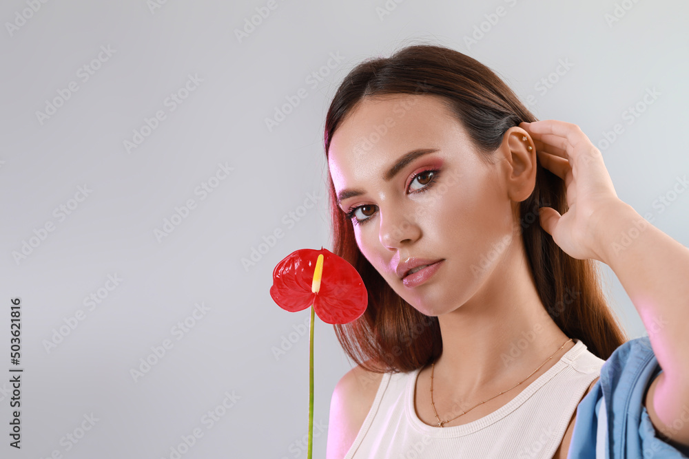 Young woman adjusting her hair and holding beautiful anthurium flower on light background