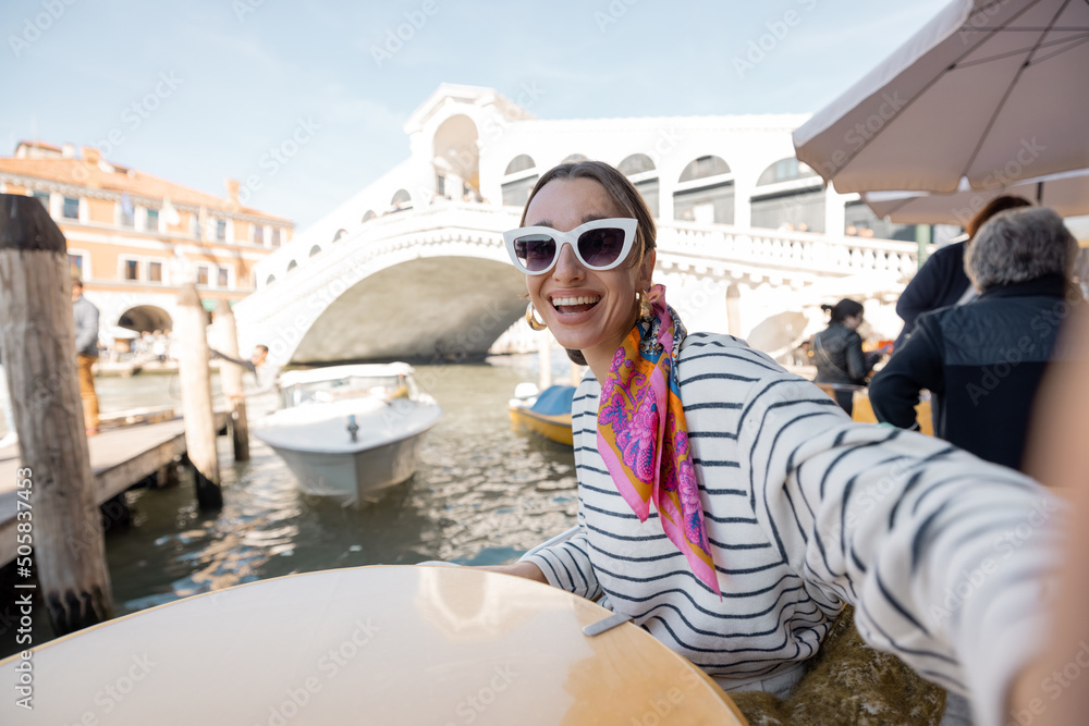 Young woman making selfie on phone while sitting at outdoor cafe near Grand Canal and Rialto bridge 