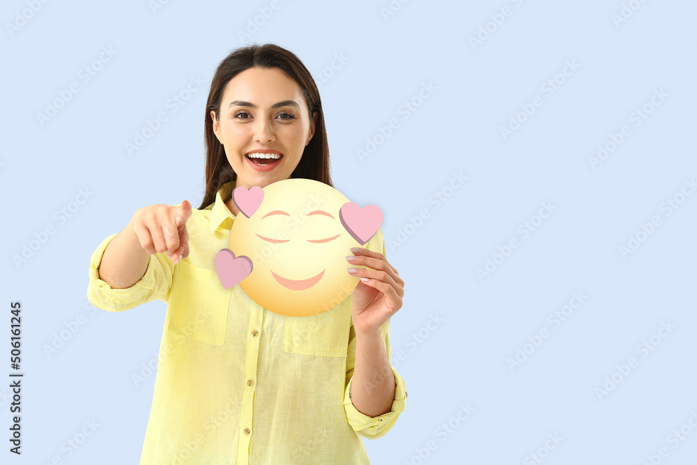 Beautiful young woman holding smiling emoticon with hearts on light blue background