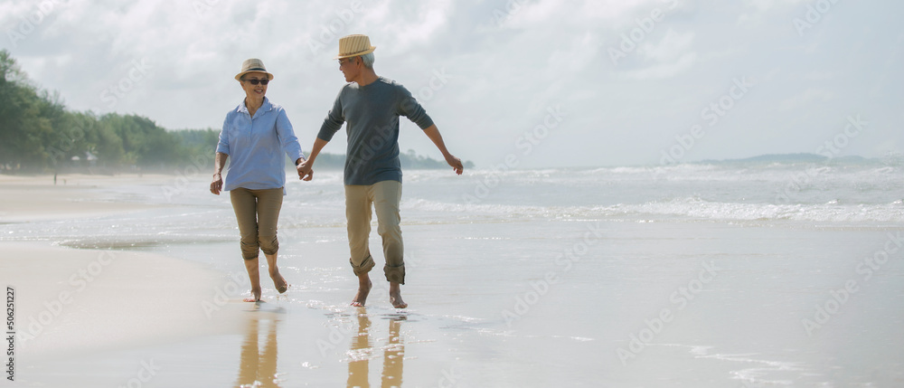 Asian senior couple wearing hat with sunglasses  holding hands  walking on the beach