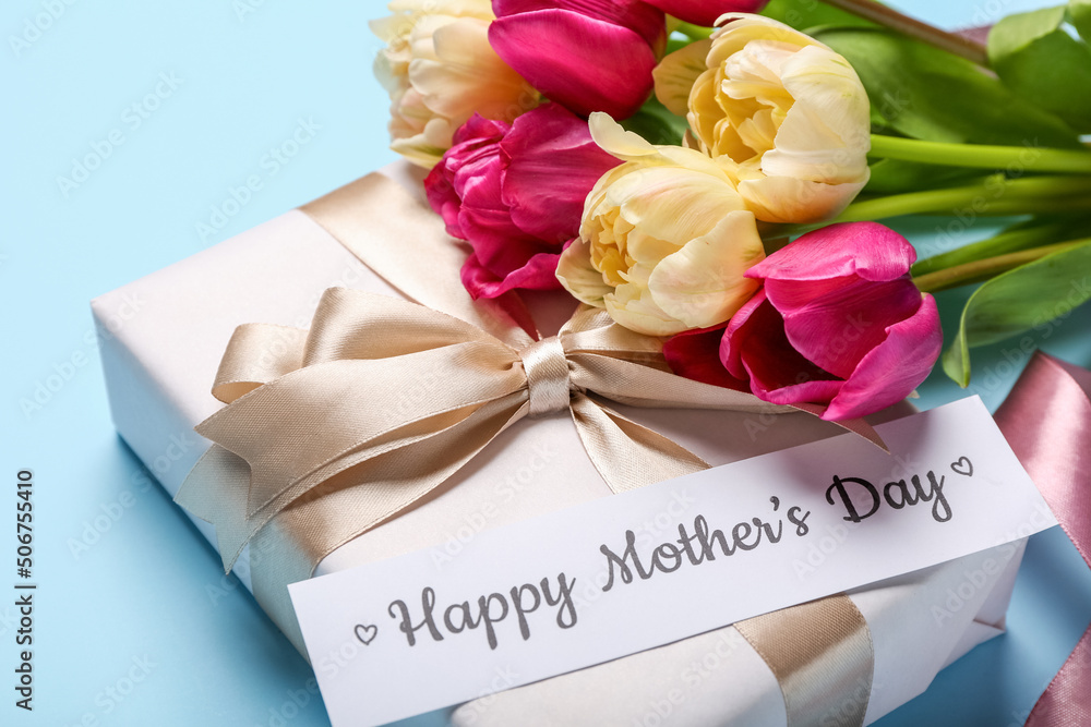 Paper with text HAPPY MOTHERS DAY, gift box and bouquet of tulips on blue background, closeup