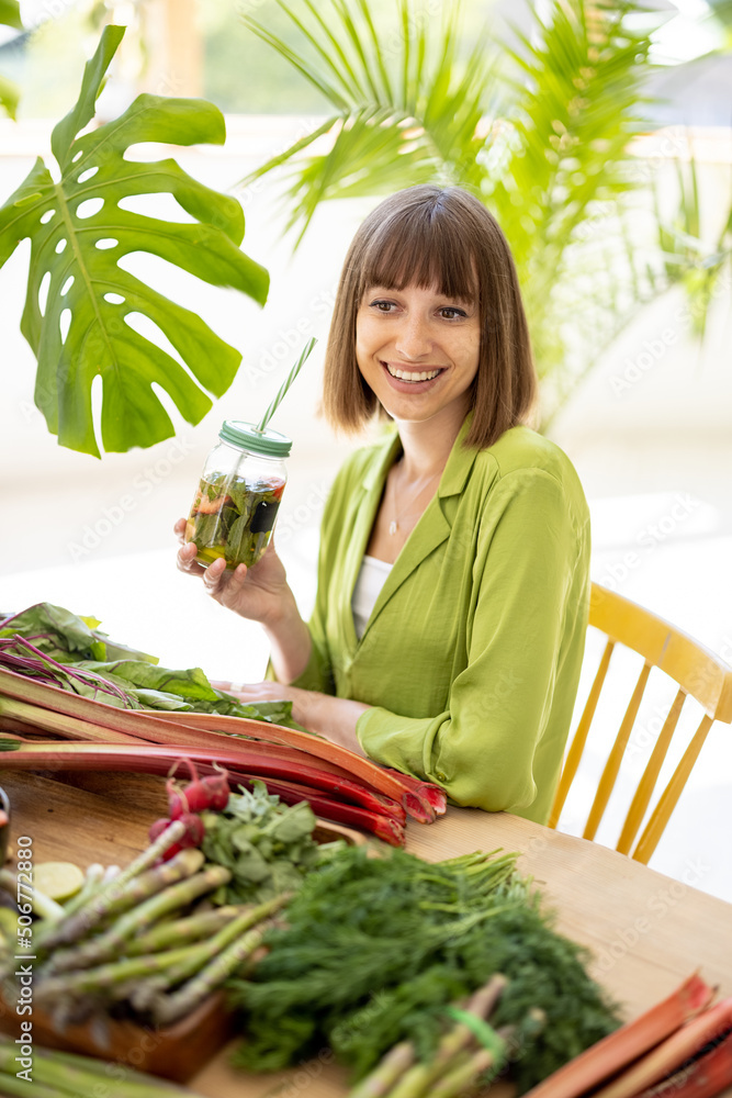 Portrait of a young woman drinks from a bottle while sitting by the table full of fresh vegetables, 