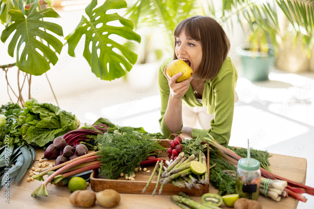 Portrait of a young cheerful woman stands by the table full of fresh vegetables, fruits and greens w