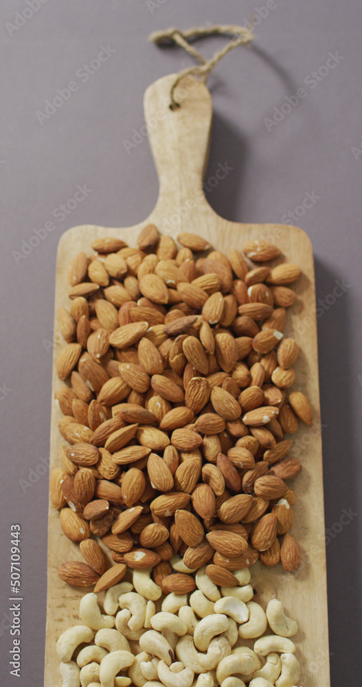 Vertical image of almond and cashew nuts piled on wooden chopping board with lilac background