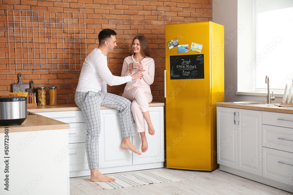 Happy young couple in pajamas drinking coffee near refrigerator in kitchen