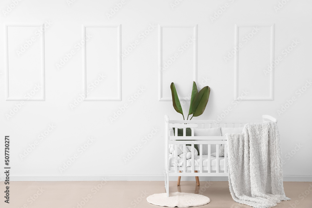 Baby crib with blanket, pillows and palm leaves near light wall