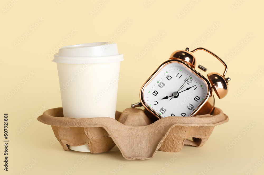 Holder with takeaway paper cup and alarm clock on beige background