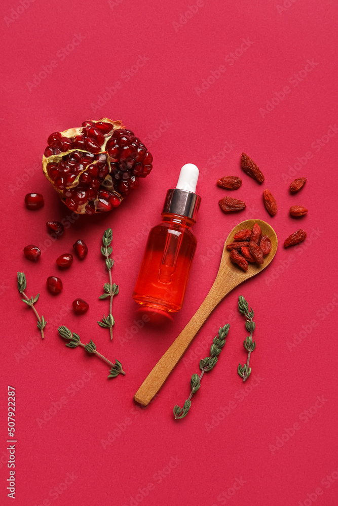 Bottle of natural serum, spoon with goji berries, thyme and pomegranate on red background