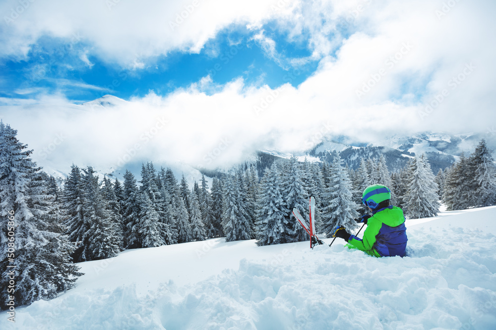 Kid sit in snow with ski over fir forest after heavy snowfall