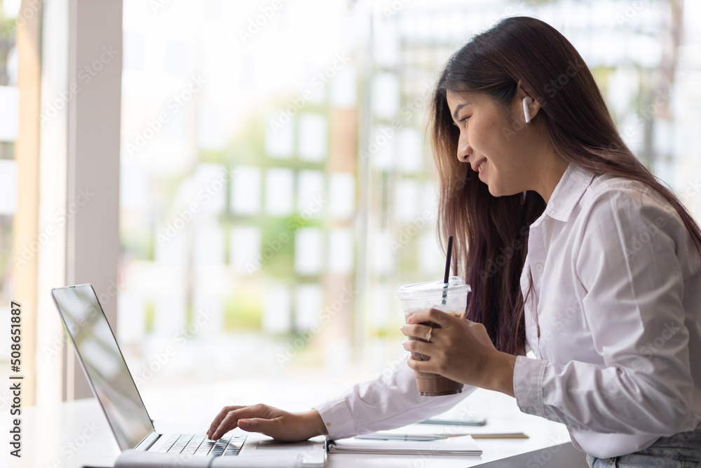 Attractive Asian secretary or businesswoman holding iced coffee sitting on laptop in office.