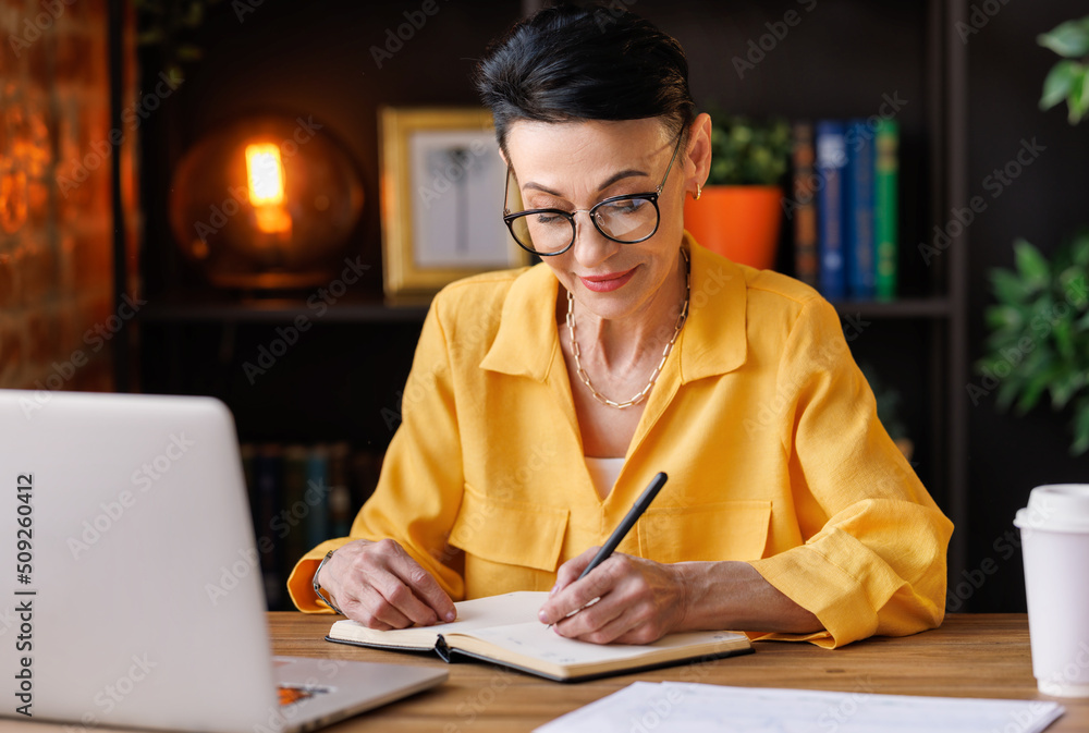 Middle aged businesswoman working at laptop,   take notes in home office