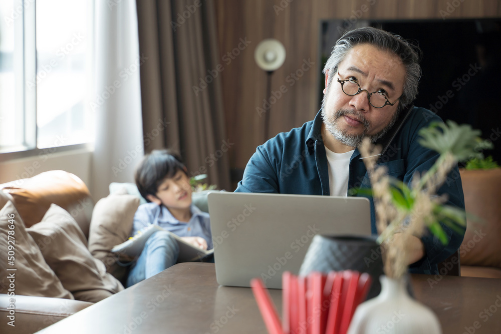 一名亚洲中年男子带着儿子在家用笔记本电脑工作，在背面画了一幅画。