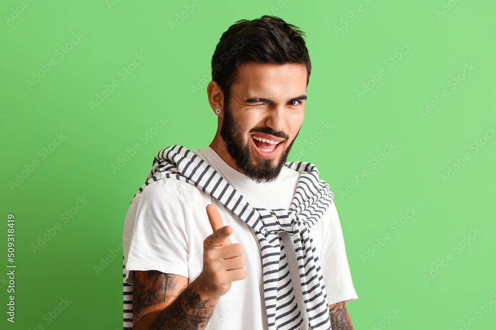 Young bearded man pointing at viewer and winking on green background