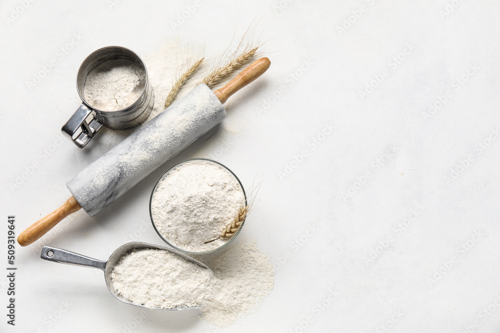 Bowl and scoop with wheat flour and rolling pin on light background