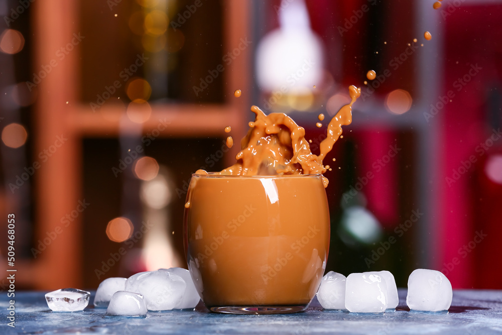 Glass of tasty iced coffee with splashes on table against blurred background