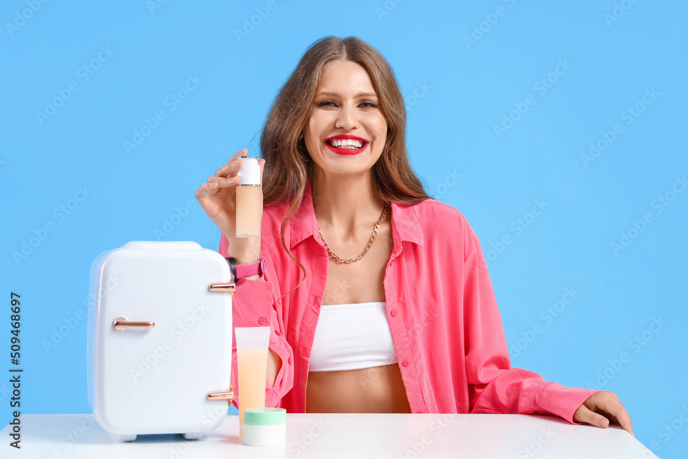 Beautiful young woman with small refrigerator for cosmetic products at table on blue background