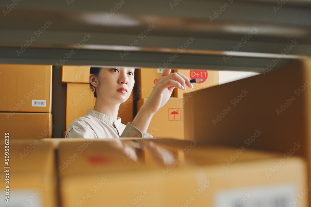 Asian woman working at online store warehouse checking inventory stock parcel boxes on shelves, onli