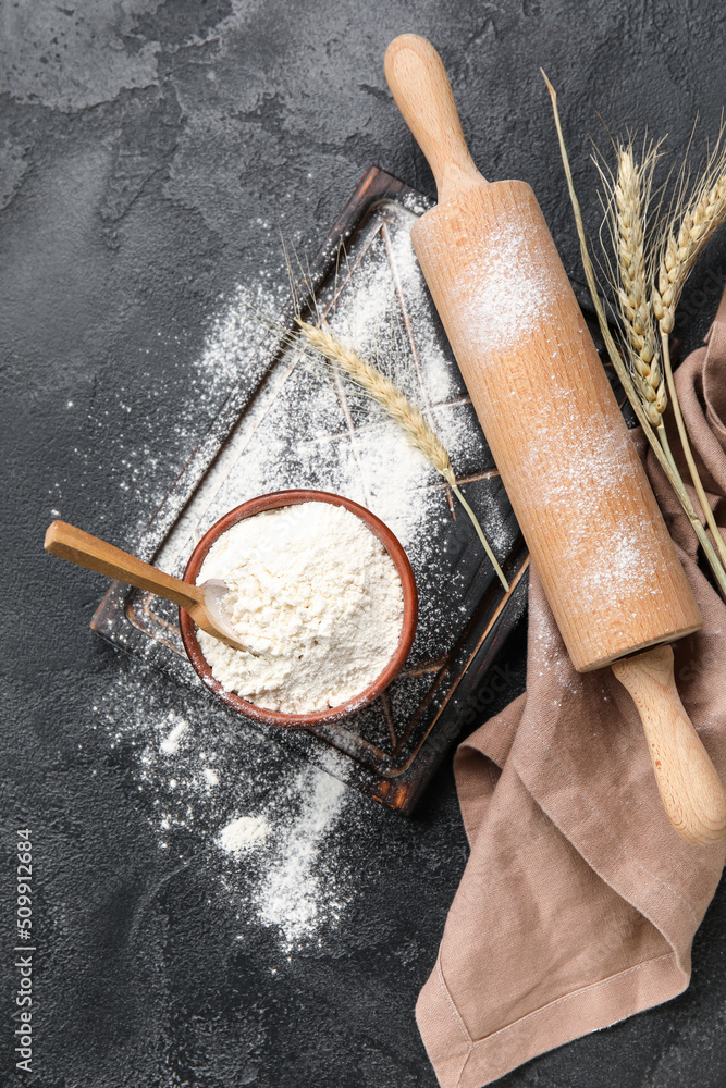 Bowl with wheat flour and wooden rolling pin on dark background