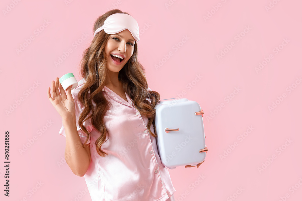 Beautiful young woman in pajamas and with small refrigerator for cosmetic products on pink backgroun