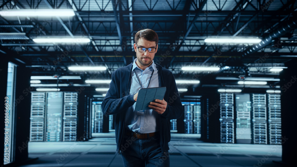 Male IT Specialist Walks Between Row of Operational Server Racks in Data Center. Engineer Uses Table