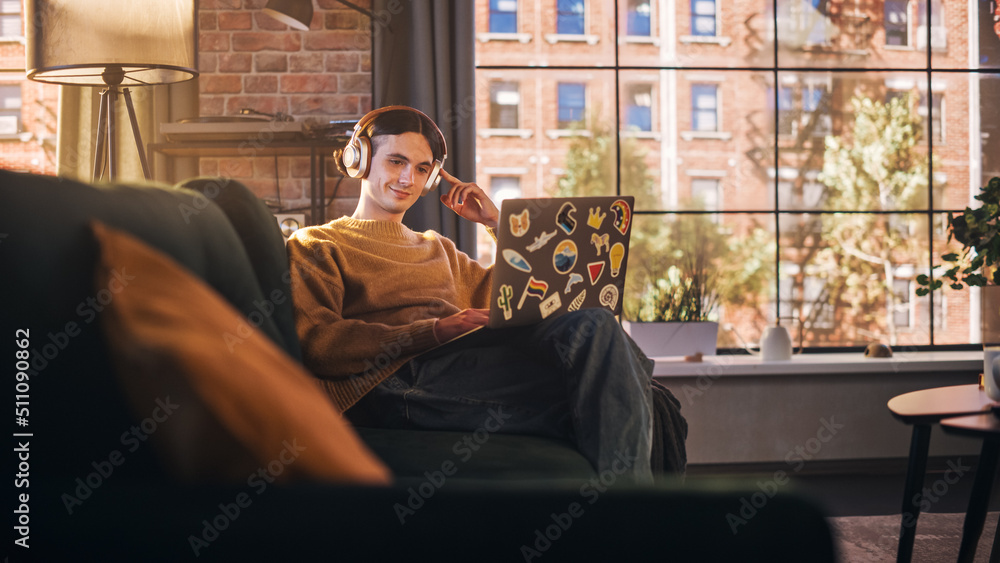 Young Handsome Man Sitting on Sofa Works on Laptop Computer in Sunny Stylish Loft Apartment. Creativ