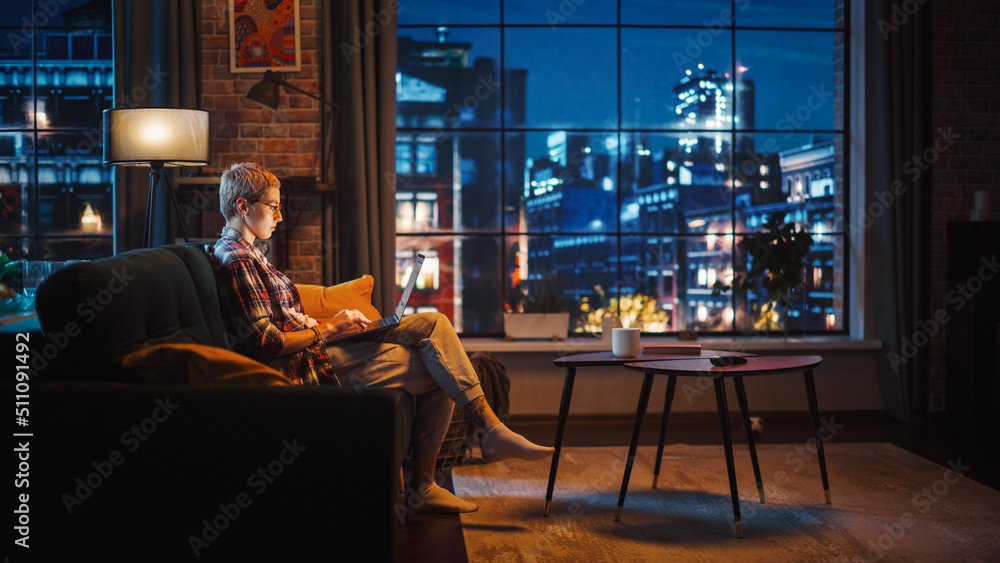 Young Beautiful Woman Sitting on Couch, Using Laptop Computer in Stylish Loft Apartment in the Eveni