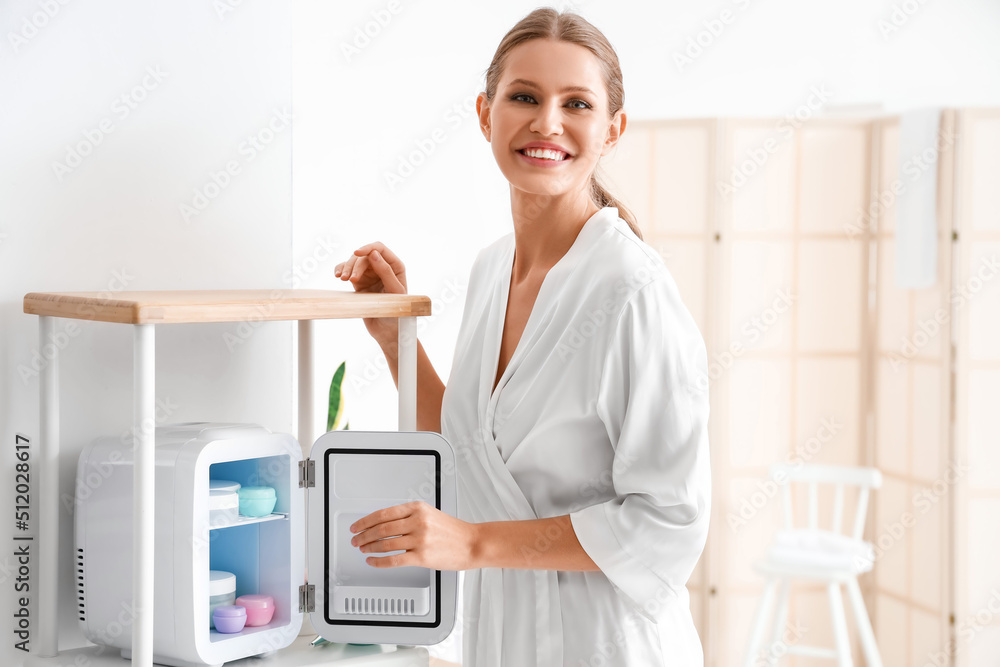 Beautiful young woman taking cream from small refrigerator for cosmetic products at home