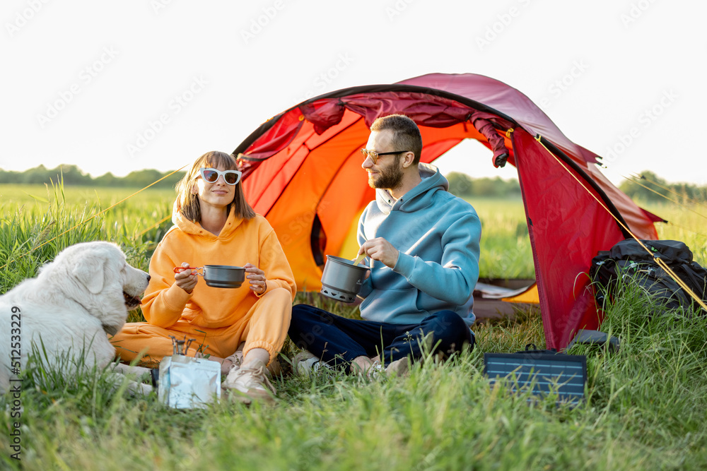 Young couple cooking food, spending summer time with a dog at campsite on the green field. Man and w