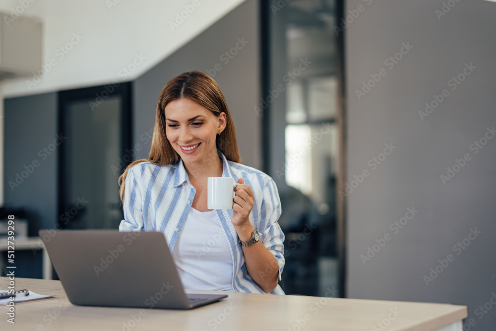 Portrait of a smiling businesswoman, holding a cup of coffee, using a laptop.