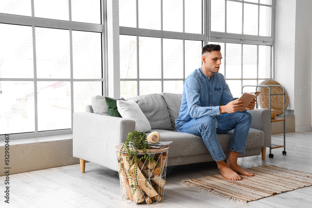 Young barefoot man reading book on sofa at home