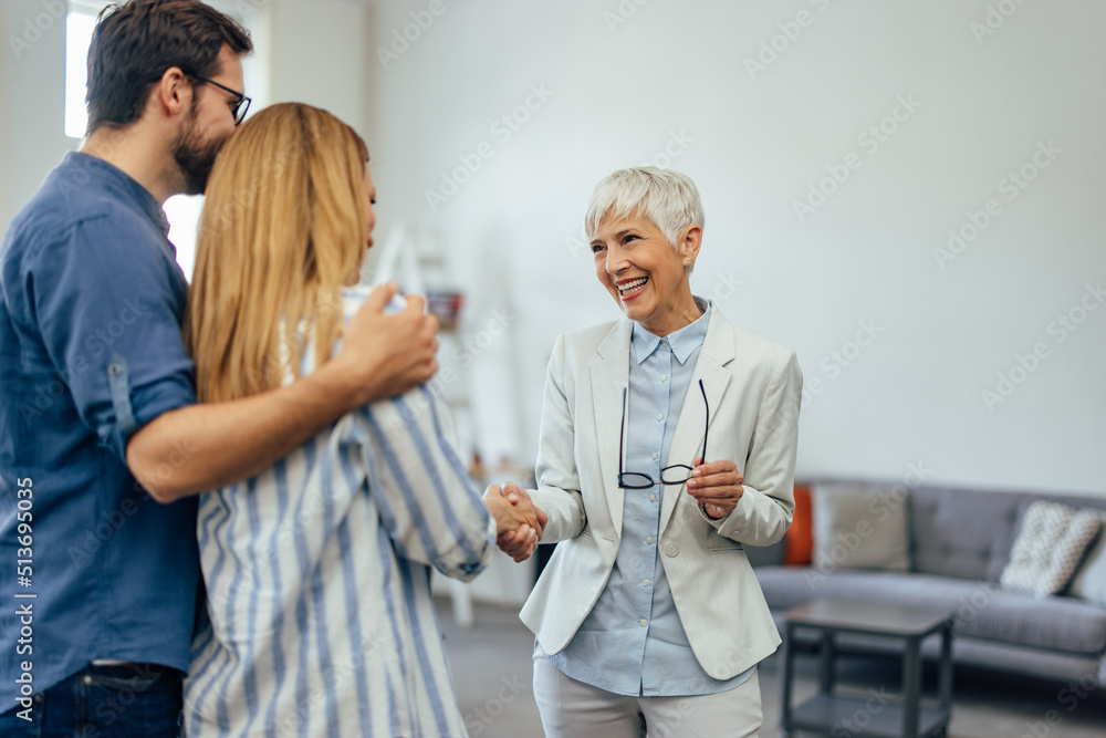 A smiling woman dressed in a suit, holding glasses, shaking hands with a young couple.