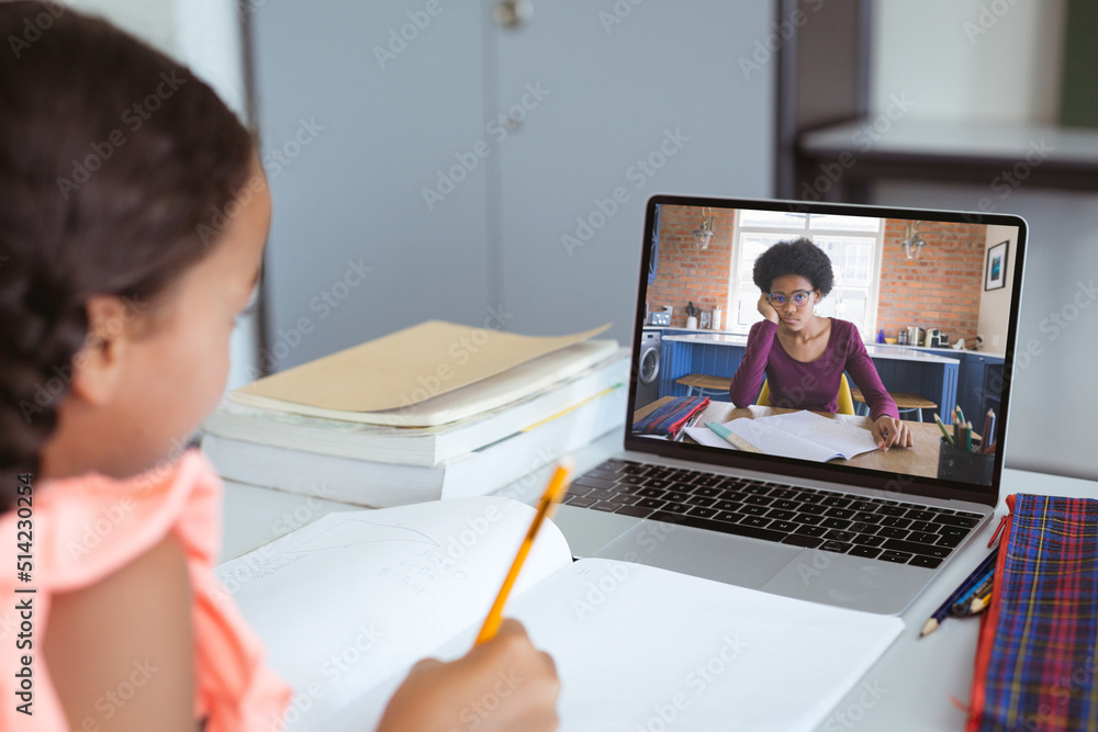 Multiracial girl writing notes while female teacher teaching over video call on laptop at home