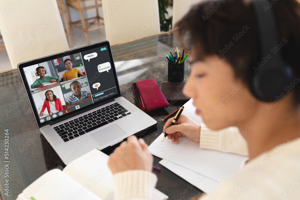 Asian teenage boy writing notes in book while attending online class over video call on laptop