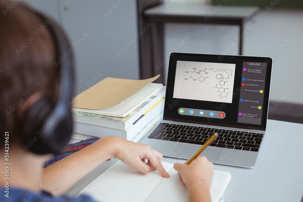 Caucasian boy writing notes in book while looking at laptop screen during online lecture at home