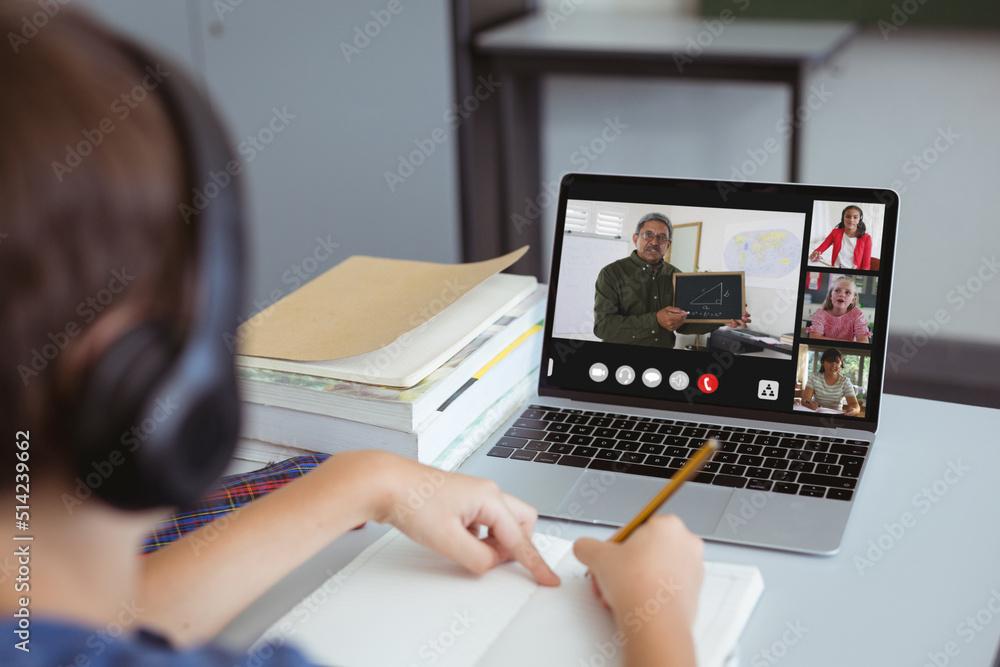 Caucasian boy wearing headphones writing in book while studying online over video call on laptop