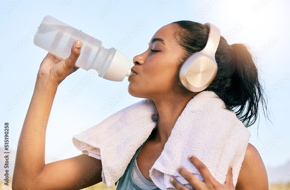 An active fit woman wearing wireless headphones drinking water from a bottle after exercising outdoo