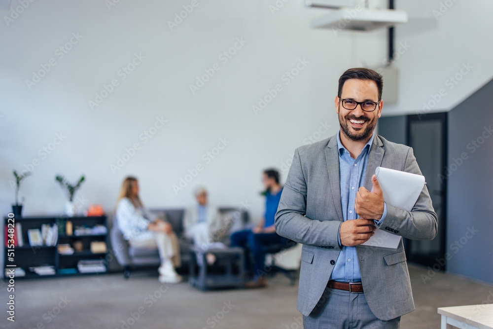 The male boss standing and posing for the photo while his colleagues sitting and talking in the back