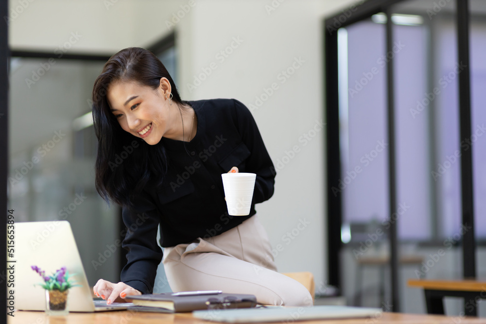 年，一位迷人的亚洲女商人在办公室里用笔记本电脑工作并喝咖啡的肖像