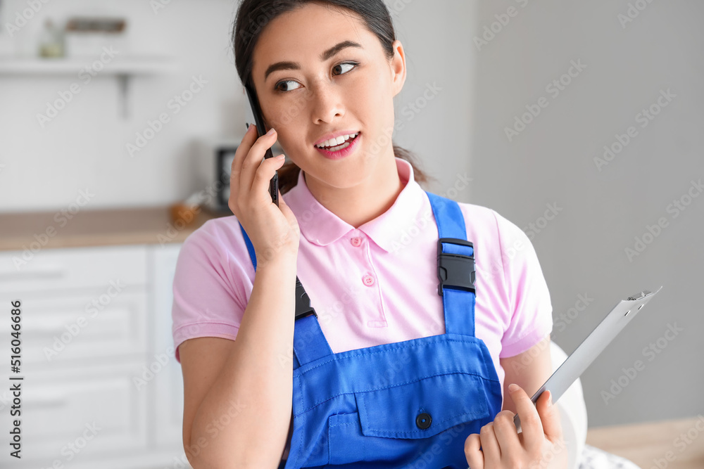 Asian female plumber with clipboard talking by mobile phone in kitchen