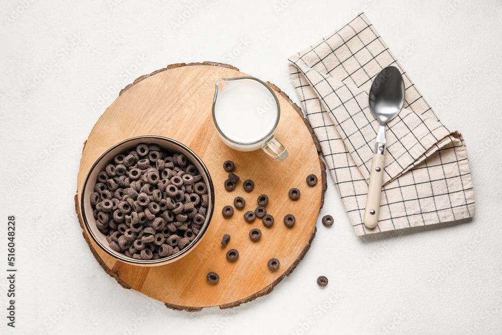 Wooden board with bowl of black cereal rings and milk on light background