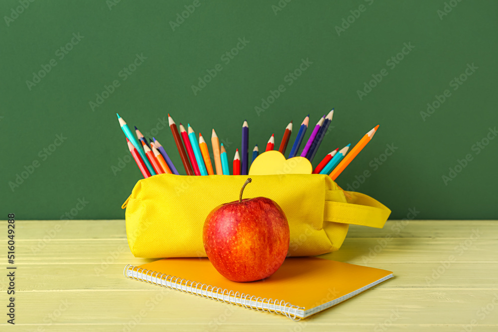 Pencil case with apple and notebook on table against green background