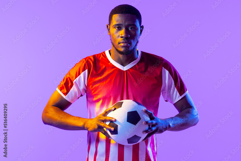 Portrait of african american male soccer player with football over neon pink lighting