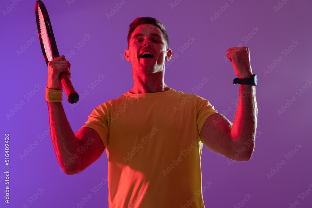 Portrait of happy caucasian male tennis player with tennis racket over neon pink lighting