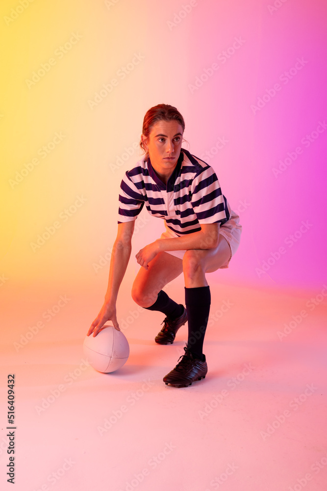 Caucasian female rugby player crouching with rugby ball over neon pink lighting