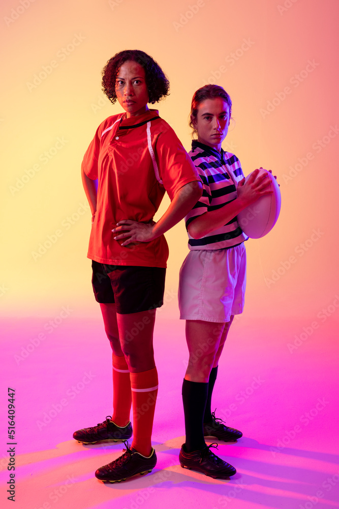 Portrait of two diverse female rugby players with rugby ball over neon pink lighting