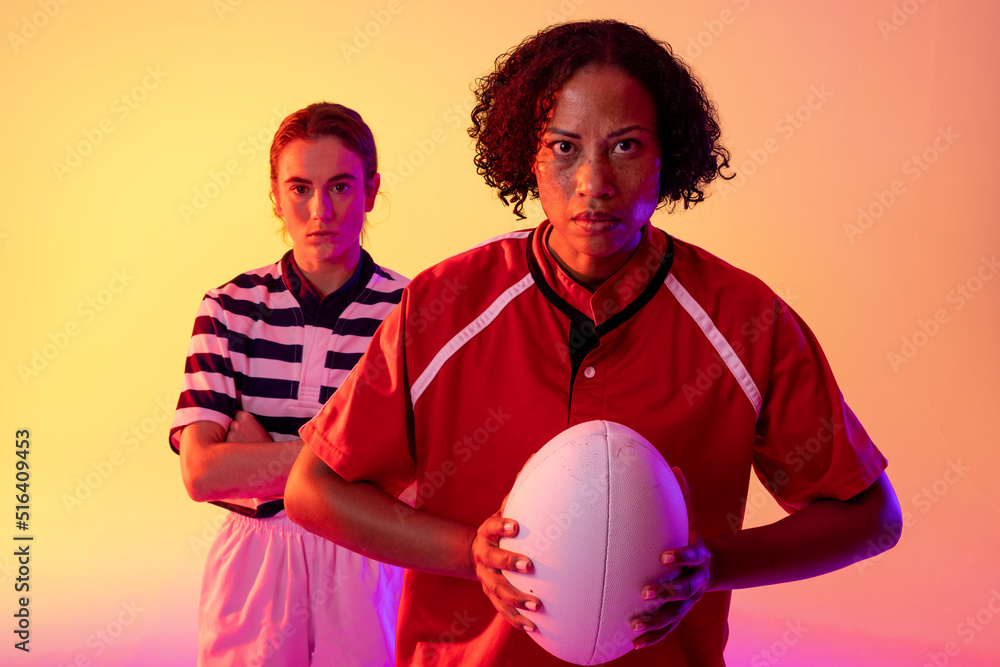 Portrait of diverse female rugby players with rugby ball over neon pink lighting