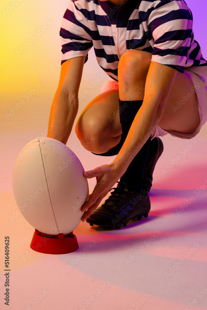 Caucasian female rugby player crouching with rugby ball over neon pink lighting